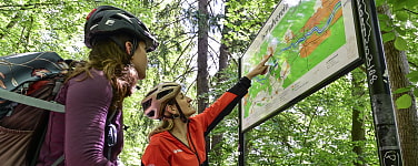 Zwei Personen mit Mountainbikes stehen vor einem Wegschild im Isartal und besprechen den Wegverlauf.